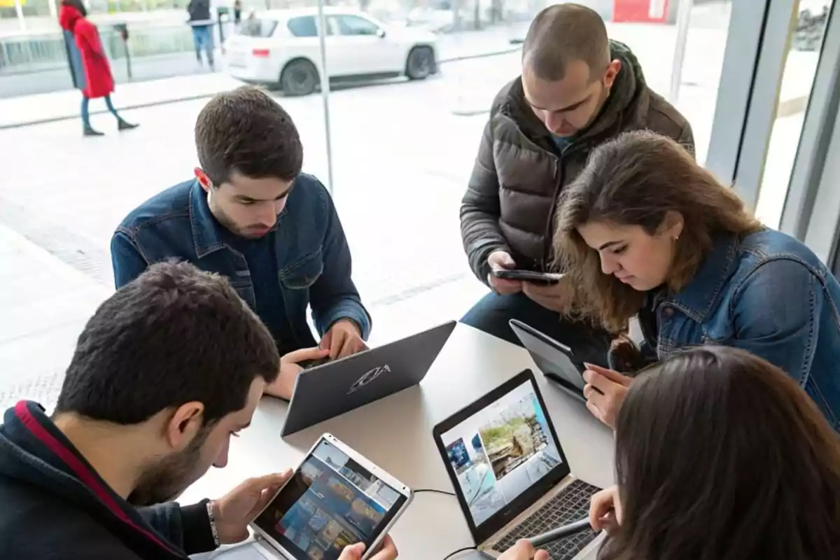 Un grupo de personas usando dispositivos electrónicos en una mesa junto a una ventana.