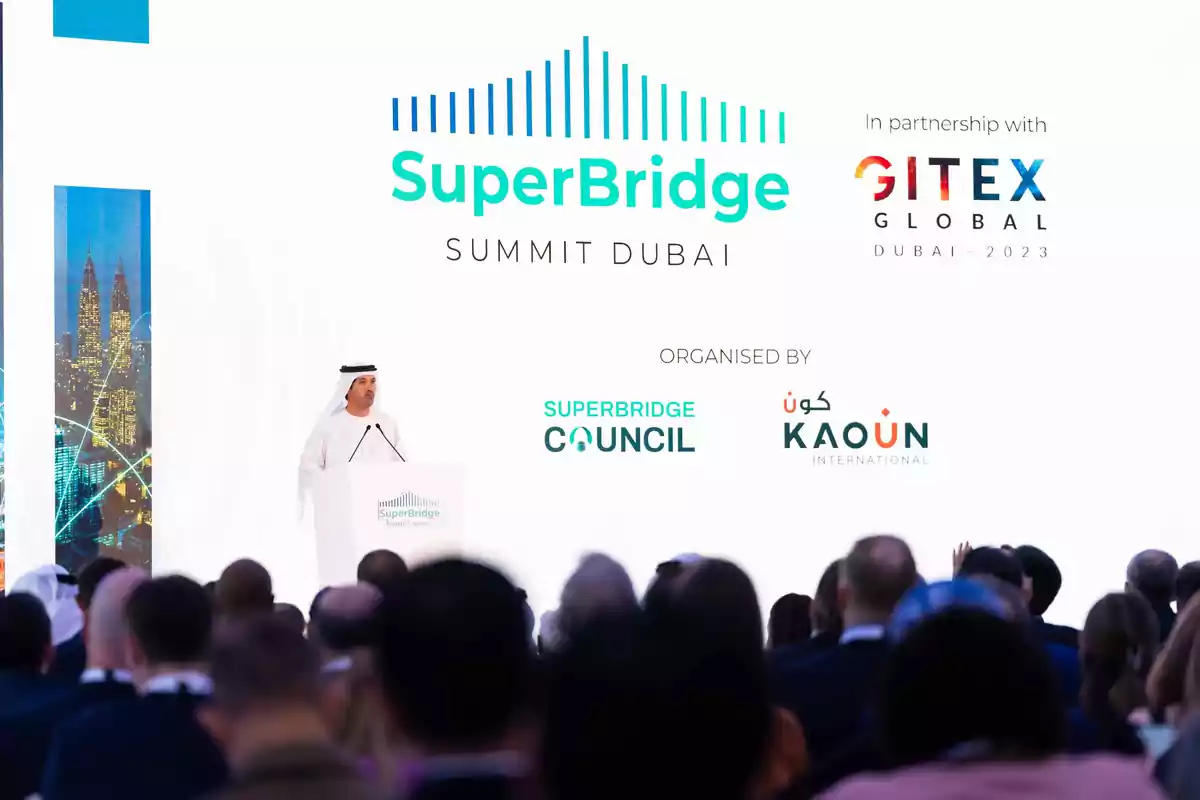 Un hombre en un podio hablando en el SuperBridge Summit Dubai con logotipos de GITEX Global, SuperBridge Council y Kaoun International en el fondo.