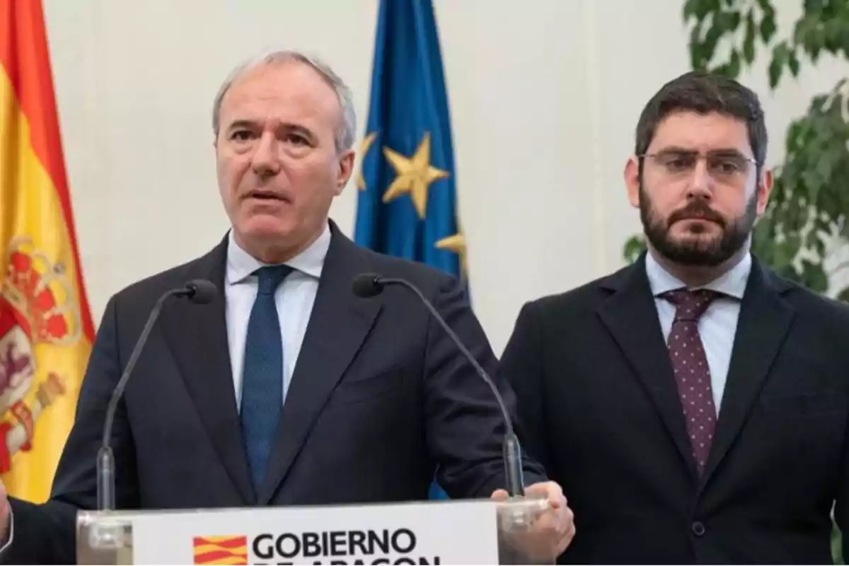 Dos hombres en traje y corbata en una conferencia de prensa con banderas de España y la Unión Europea de fondo, con un cartel que dice "Gobierno de Aragón".