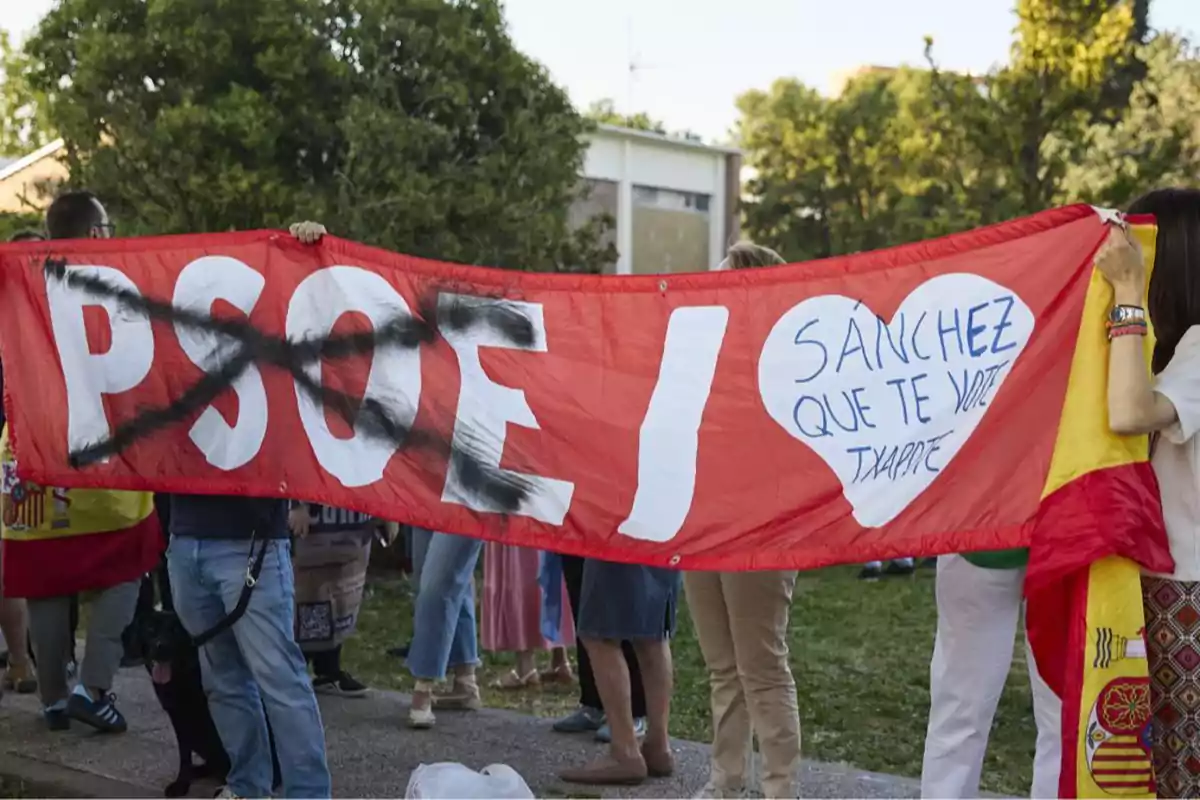 Personas sostienen una pancarta roja con las letras PSOE tachadas en negro y un corazón blanco con el mensaje Sánchez que te vote Txapote, mientras una persona sostiene una bandera de España. Personas sostienen una pancarta roja con las letras PSOE tachadas en negro y un corazón blanco con el mensaje Sánchez que te vote Txapote, mientras una persona sostiene una bandera de España.