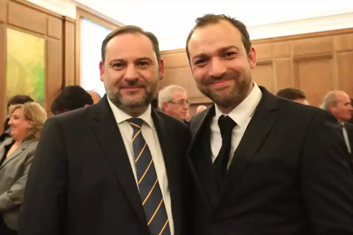 Two men in suits smile at a social event with several people in the background.