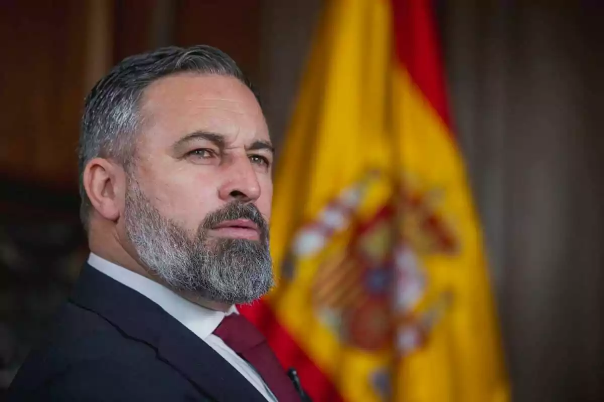 Un hombre con barba y traje oscuro está de perfil frente a una bandera de España.