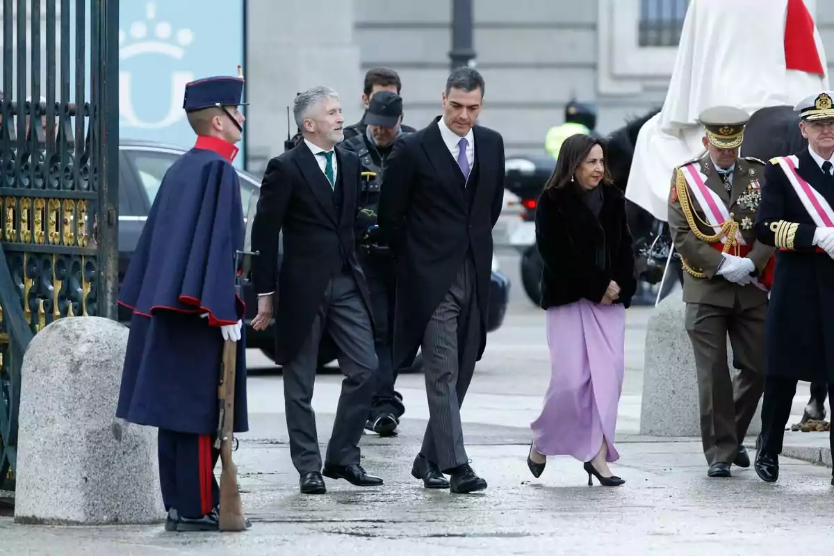 Un grupo de personas vestidas formalmente camina junto a un guardia y oficiales militares en un evento al aire libre.