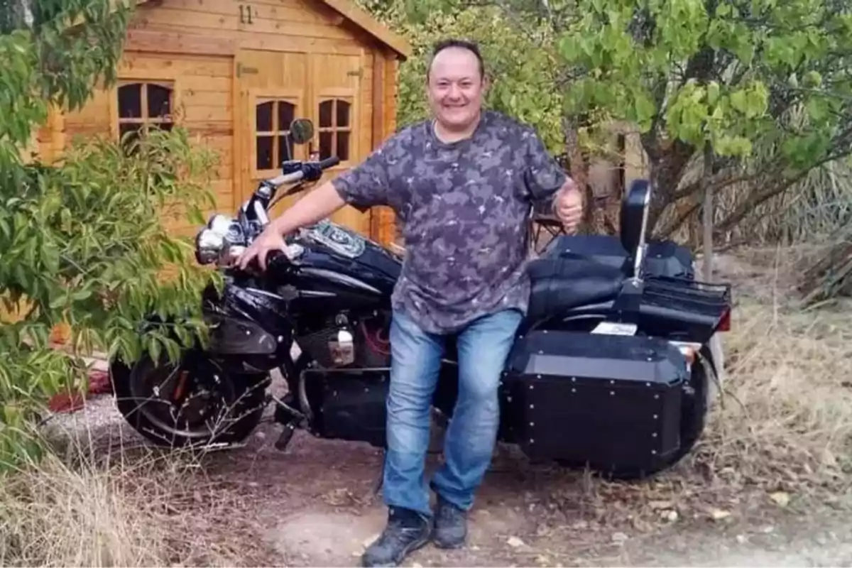 Hombre sonriendo junto a una motocicleta negra frente a una cabaña de madera en un entorno natural.