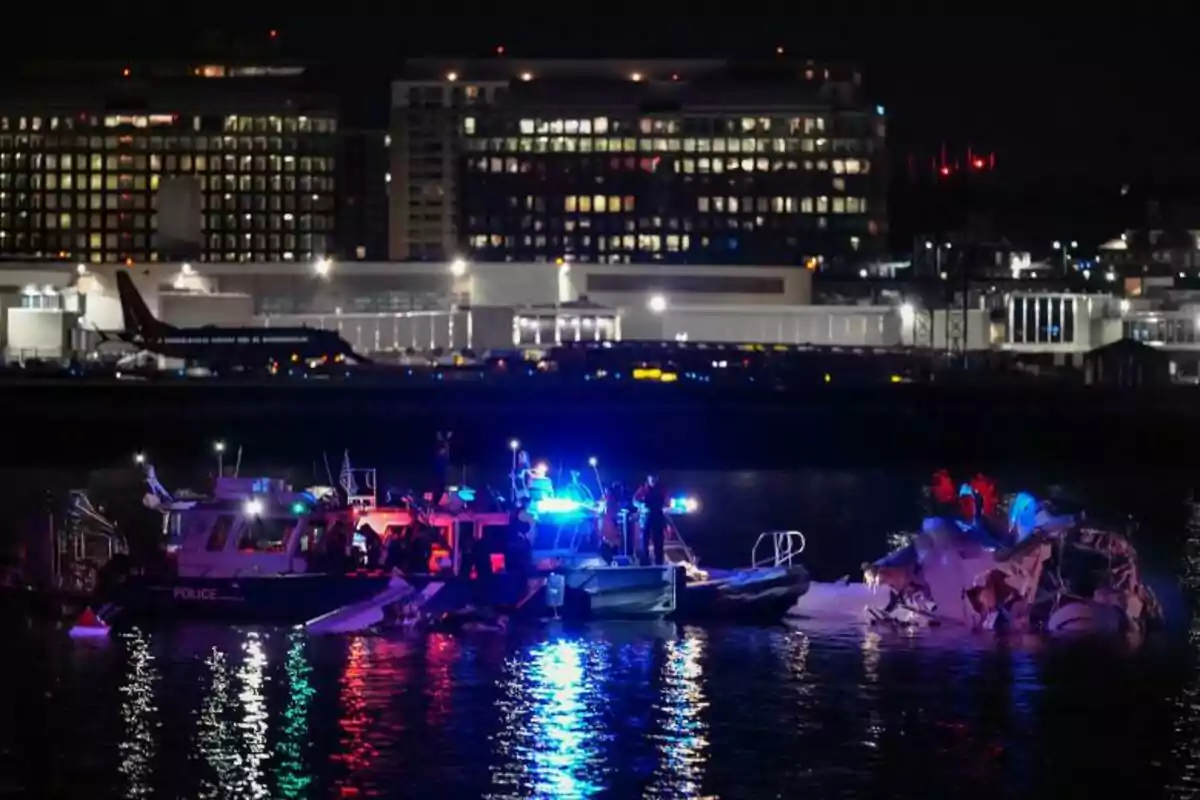 Una escena nocturna muestra un operativo de rescate en el agua con luces de emergencia brillando, mientras un edificio iluminado se ve al fondo.