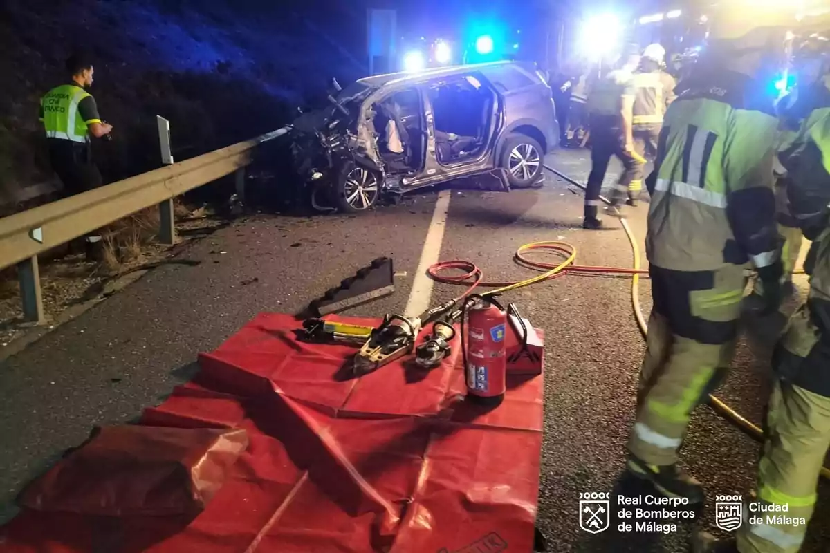 Bomberos y personal de emergencia atienden un accidente de tráfico nocturno en una carretera, con un coche gravemente dañado junto a la valla de seguridad y herramientas de rescate sobre una lona roja en primer plano