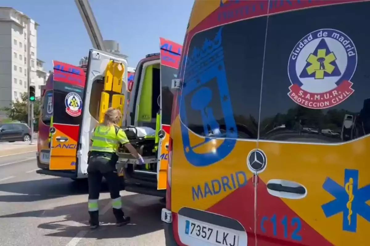 Una trabajadora de emergencias médicas de SAMUR Protección Civil de Madrid está manipulando una camilla junto a dos ambulancias estacionadas en una calle.