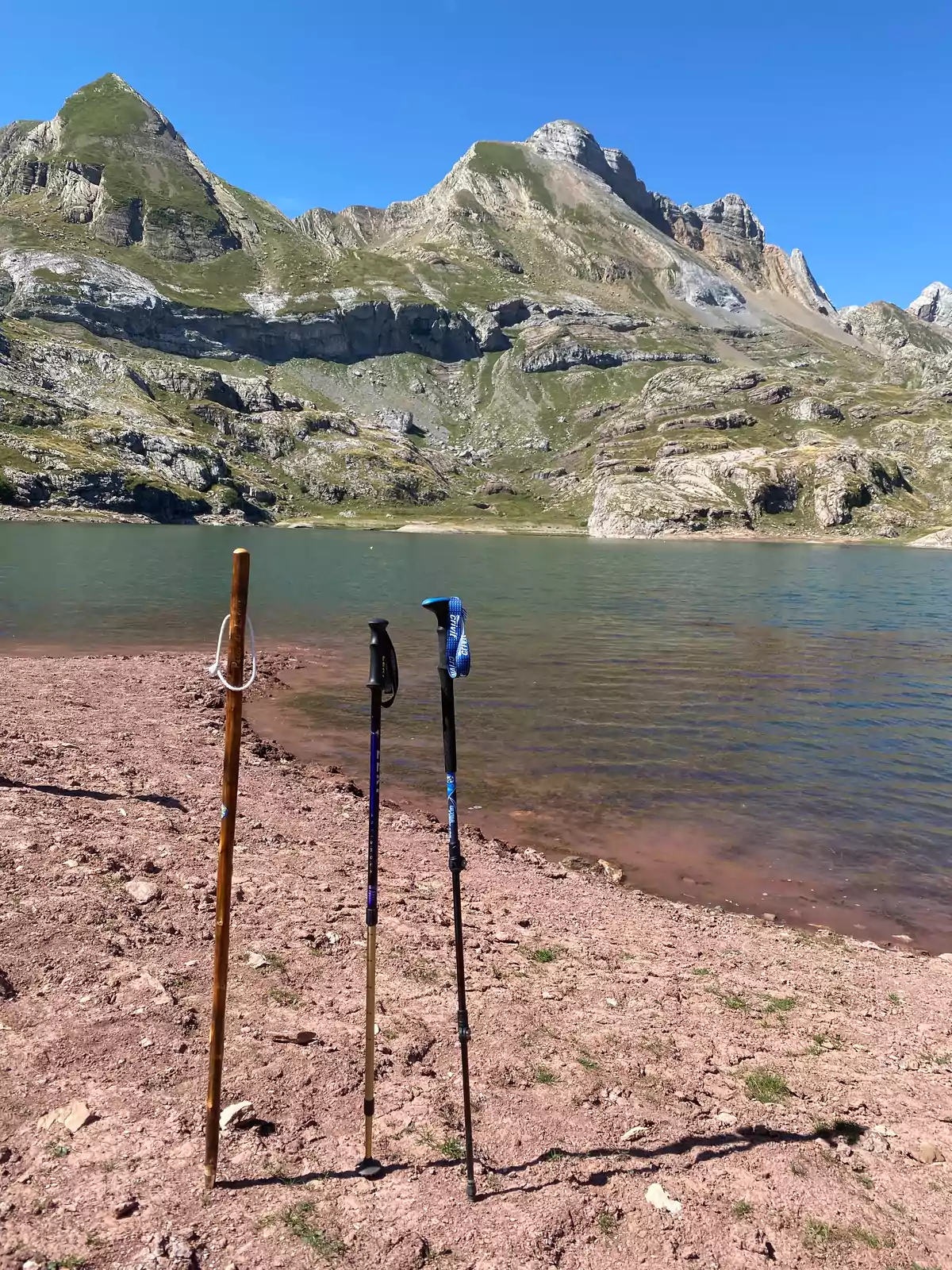Tres bastones de senderismo clavados en la orilla de un lago con montañas al fondo bajo un cielo despejado. Tres bastones de senderismo clavados en la orilla de un lago con montañas al fondo bajo un cielo despejado.