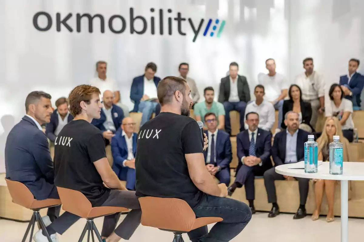 Personas en una conferencia de prensa de OK Mobility, con dos hombres de espaldas usando camisetas de LIUX y una audiencia sentada al fondo.