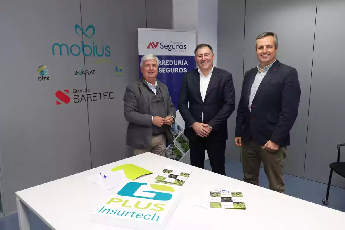 Tres hombres de pie junto a una mesa blanca con documentos y un cartel que dice "PLUS Insurtech" en una oficina con logotipos de empresas en la pared.