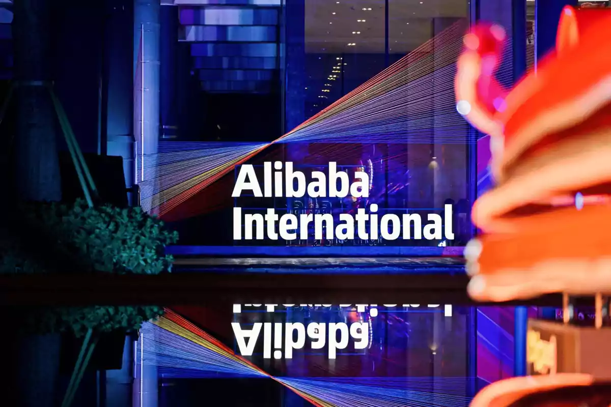 Un letrero iluminado de Alibaba International se refleja en una superficie brillante frente a un edificio moderno.