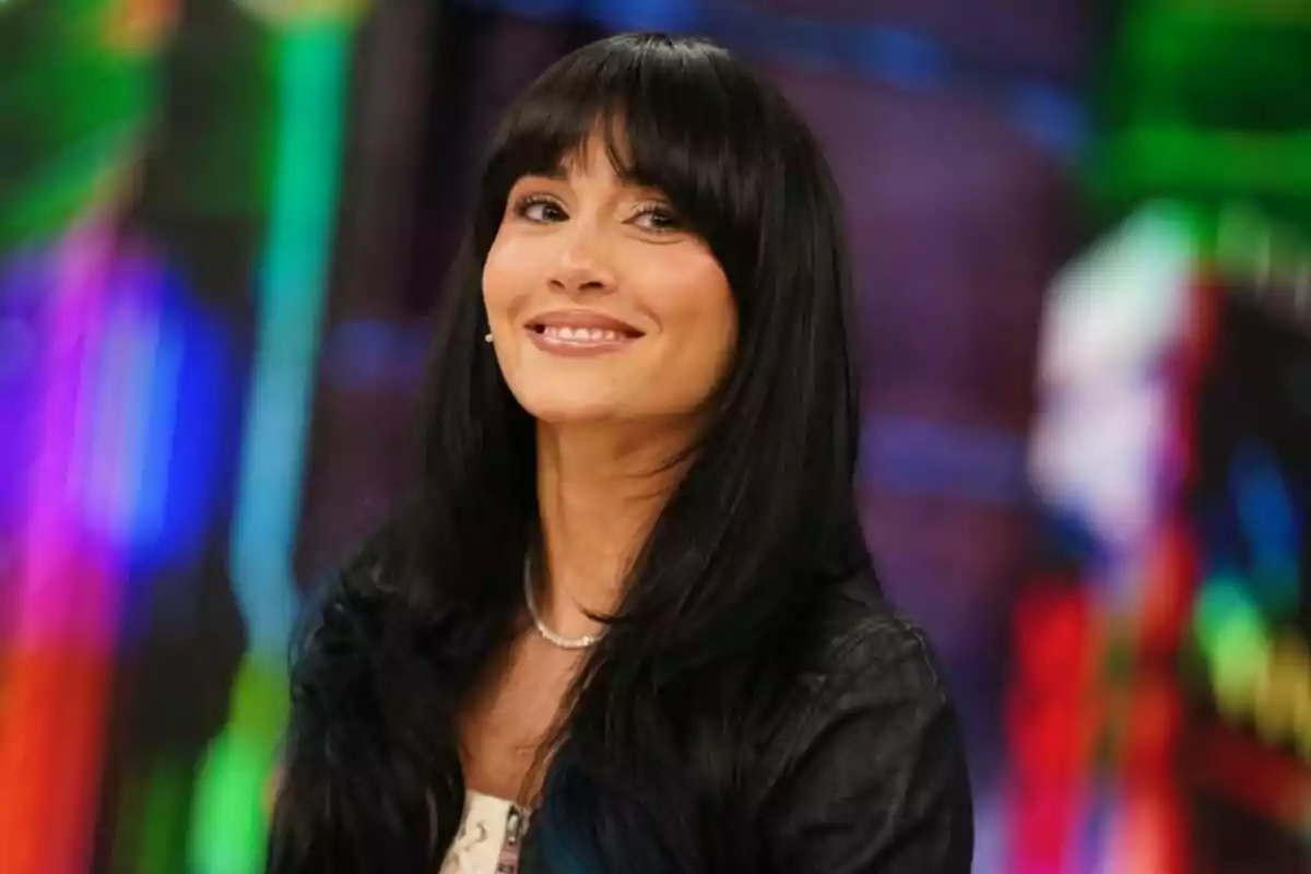 Aitana con cabello largo y oscuro con flequillo sonriendo frente a un fondo colorido y desenfocado en 'El Hormiguero'. Aitana con cabello largo y oscuro con flequillo sonriendo frente a un fondo colorido y desenfocado en 'El Hormiguero'.