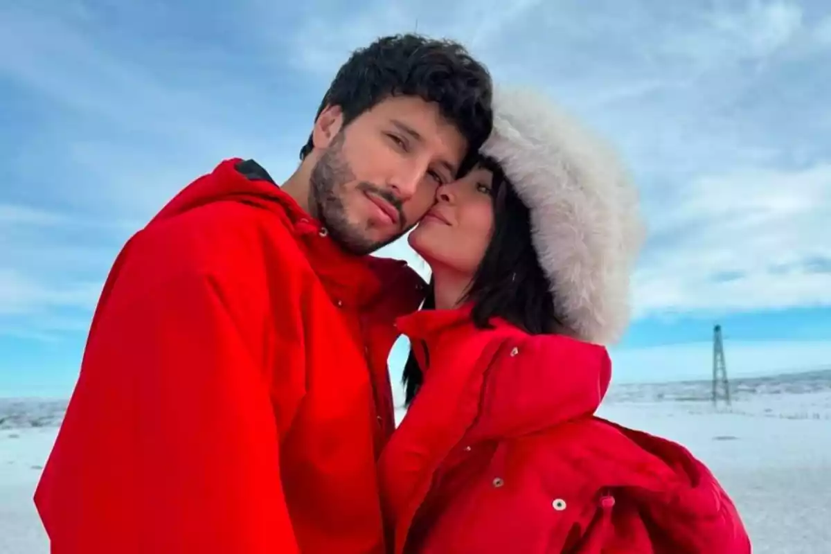 Aitana y Sebastián Yatra posan en medio de un paisaje nevado, ambos ataviados con abrigos rojos y Aitana luciendo un sombrero de pelo sintético blanco