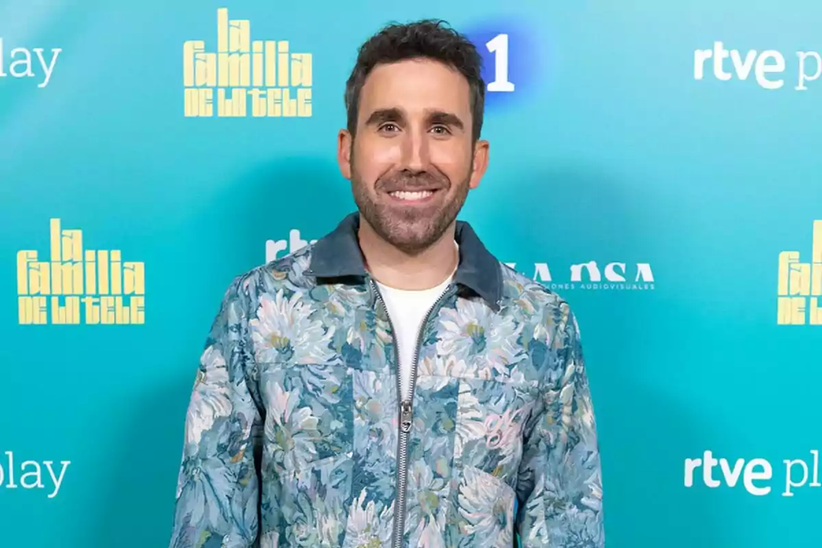 Aitor Albizua sonriente con barba y cabello oscuro lleva una chaqueta con estampado floral sobre una camiseta blanca, posando frente a un fondo azul con logotipos de Aitor Albizua sonriente con barba y cabello oscuro lleva una chaqueta con estampado floral sobre una camiseta blanca, posando frente a un fondo azul con logotipos de