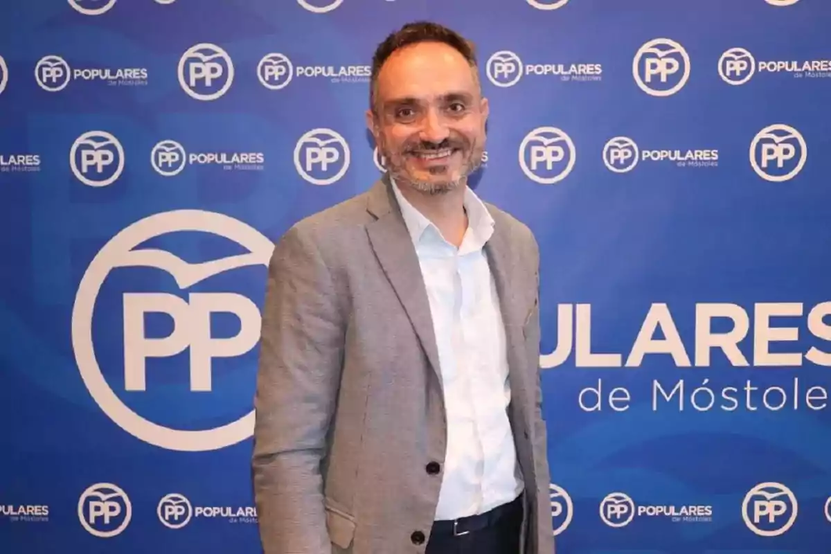 Hombre sonriendo frente a un fondo azul con logotipos del Partido Popular de Móstoles.