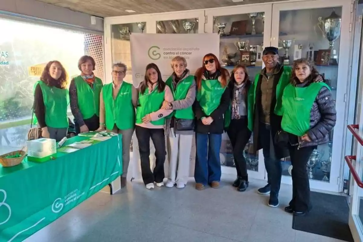 A group of people in green vests pose in front of a table and a Spanish Association Against Cancer sign in an indoor space.