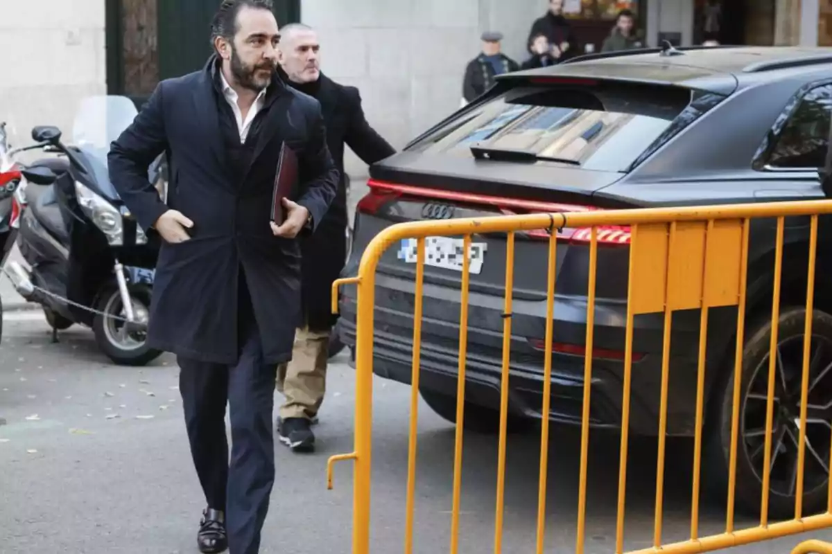 Un hombre con abrigo oscuro camina por la calle junto a un coche negro y una barrera amarilla. Un hombre con abrigo oscuro camina por la calle junto a un coche negro y una barrera amarilla.