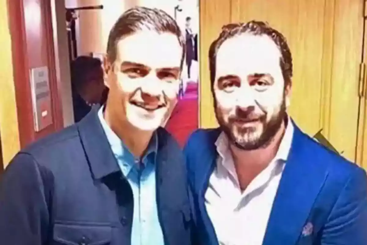 Dos hombres posan juntos para una foto en un interior con puertas de madera al fondo.