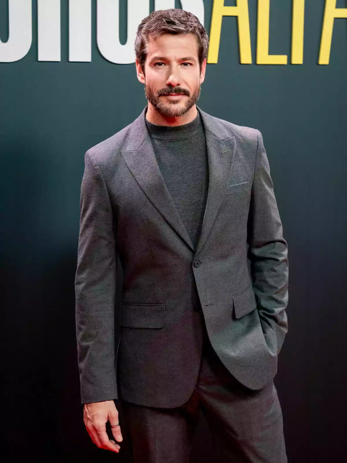 Alejandro Albarracín con barba y cabello oscuro vistiendo un traje gris oscuro posando frente a un fondo con letras grandes.