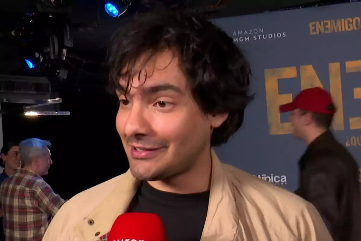 Alejandro Reyes con chaqueta beige sostiene un micrófono rojo en un evento con un fondo azul que tiene texto y logotipos. Alejandro Reyes con chaqueta beige sostiene un micrófono rojo en un evento con un fondo azul que tiene texto y logotipos.