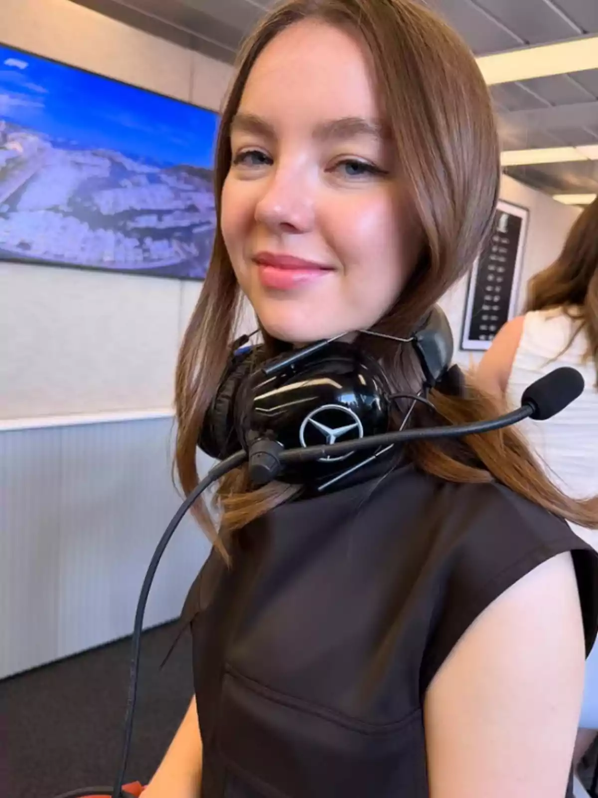 A person with headphones and a microphone is in an indoor setting, with a screen on the wall displaying an image of a coastal landscape.