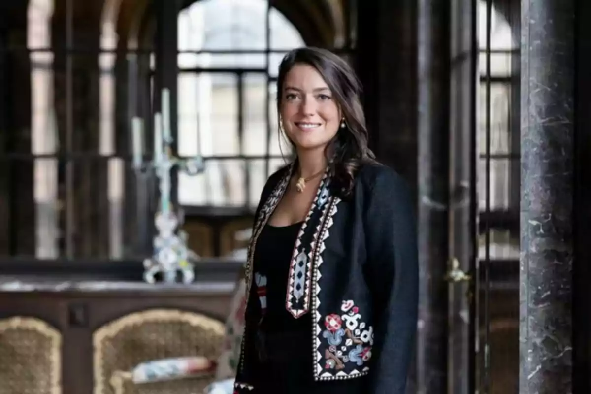 A smiling woman with dark, loose hair, dressed in a black outfit adorned with colorful embroidery, standing in an elegant interior with columns and a chandelier in the background. A smiling woman with dark, loose hair, dressed in a black outfit adorned with colorful embroidery, standing in an elegant interior with columns and a chandelier in the background.