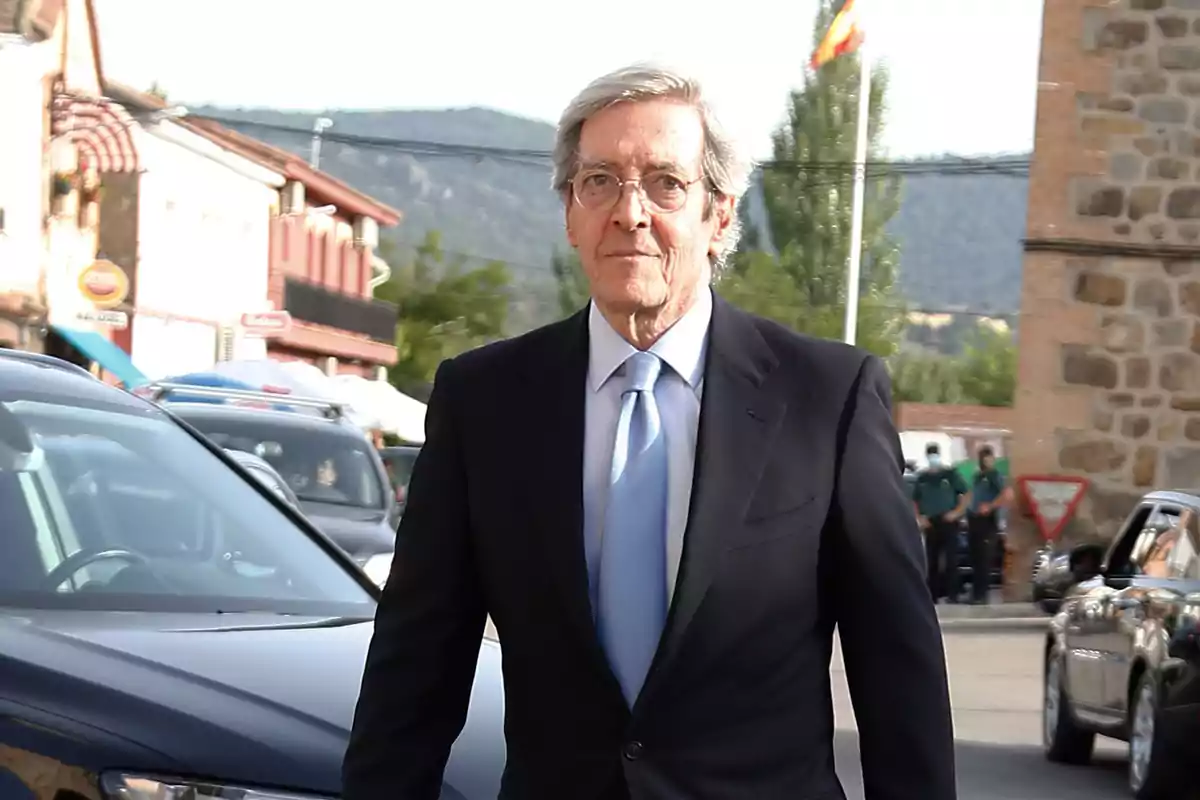 Un hombre mayor con traje oscuro y corbata azul caminando por una calle con edificios y coches al fondo. Un hombre mayor con traje oscuro y corbata azul caminando por una calle con edificios y coches al fondo.