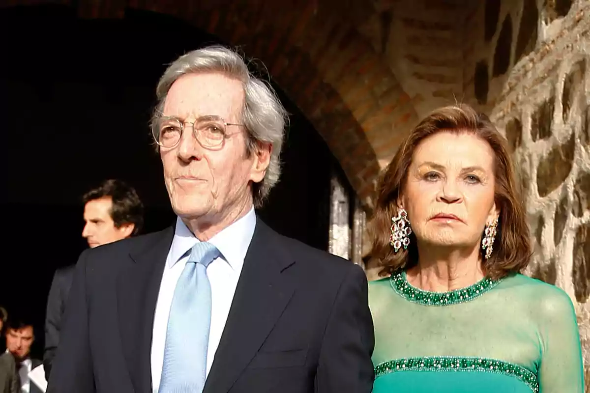Una pareja mayor elegantemente vestida, el hombre lleva traje y corbata azul claro y la mujer lleva un vestido verde con joyas llamativas, ambos están de pie frente a un fondo de piedra. Una pareja mayor elegantemente vestida, el hombre lleva traje y corbata azul claro y la mujer lleva un vestido verde con joyas llamativas, ambos están de pie frente a un fondo de piedra.