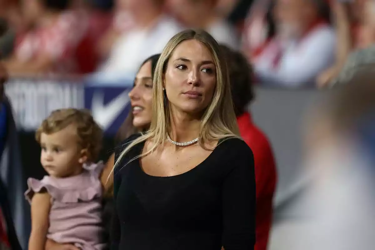 Alice Campello con un collar de diamantes y una camiseta negra está de pie en un evento deportivo, mientras una mujer detrás de ella sostiene a una niña pequeña.