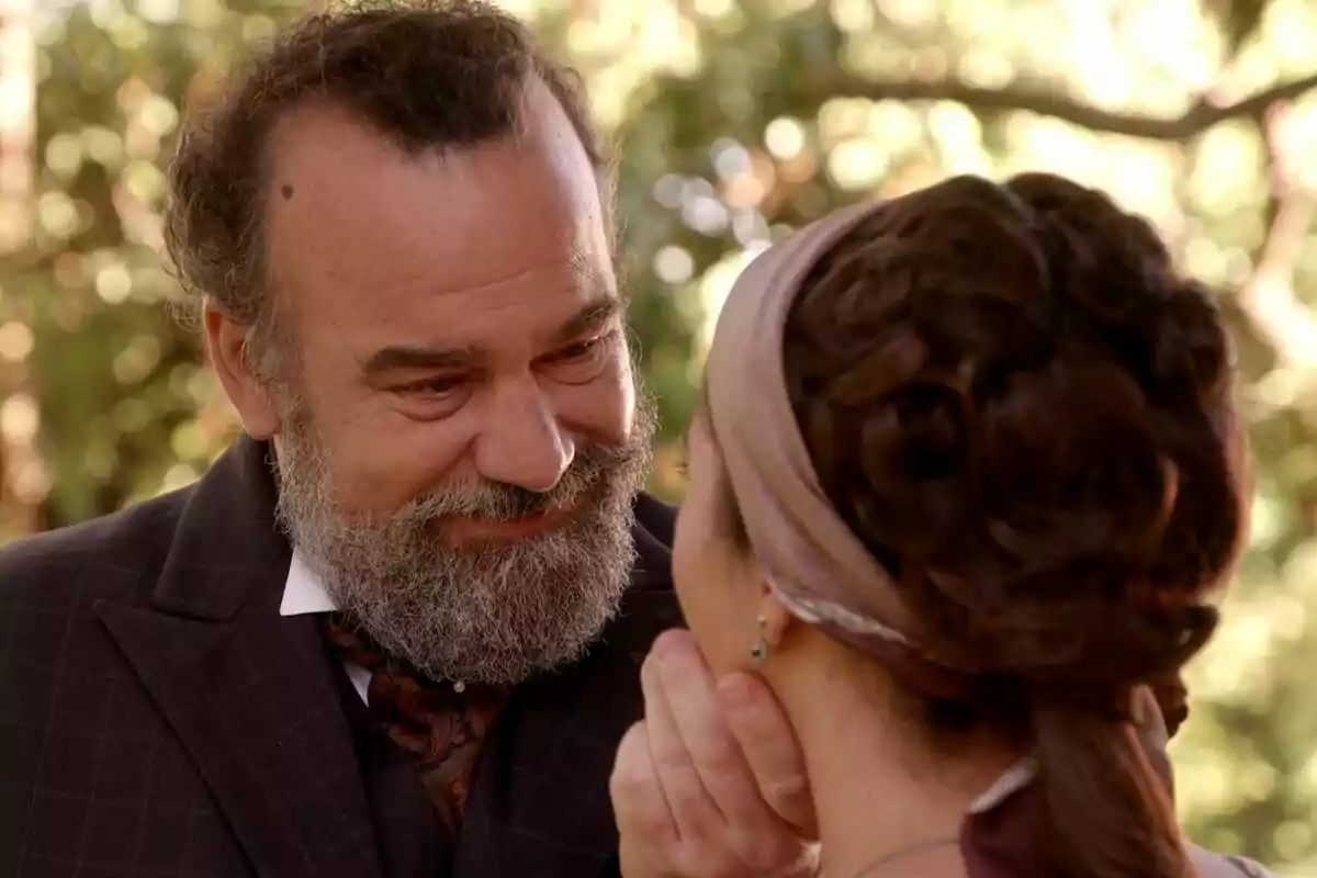 Manuel Regueiro con barba sonríe mientras sostiene el rostro de Carmen Asecas con un pañuelo en el cabello en un entorno al aire libre en La Promesa.