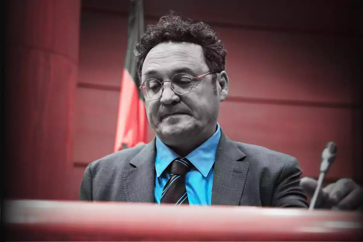 Un hombre con gafas y traje azul está de pie frente a un micrófono con una expresión seria, con un fondo rojo y una bandera detrás.