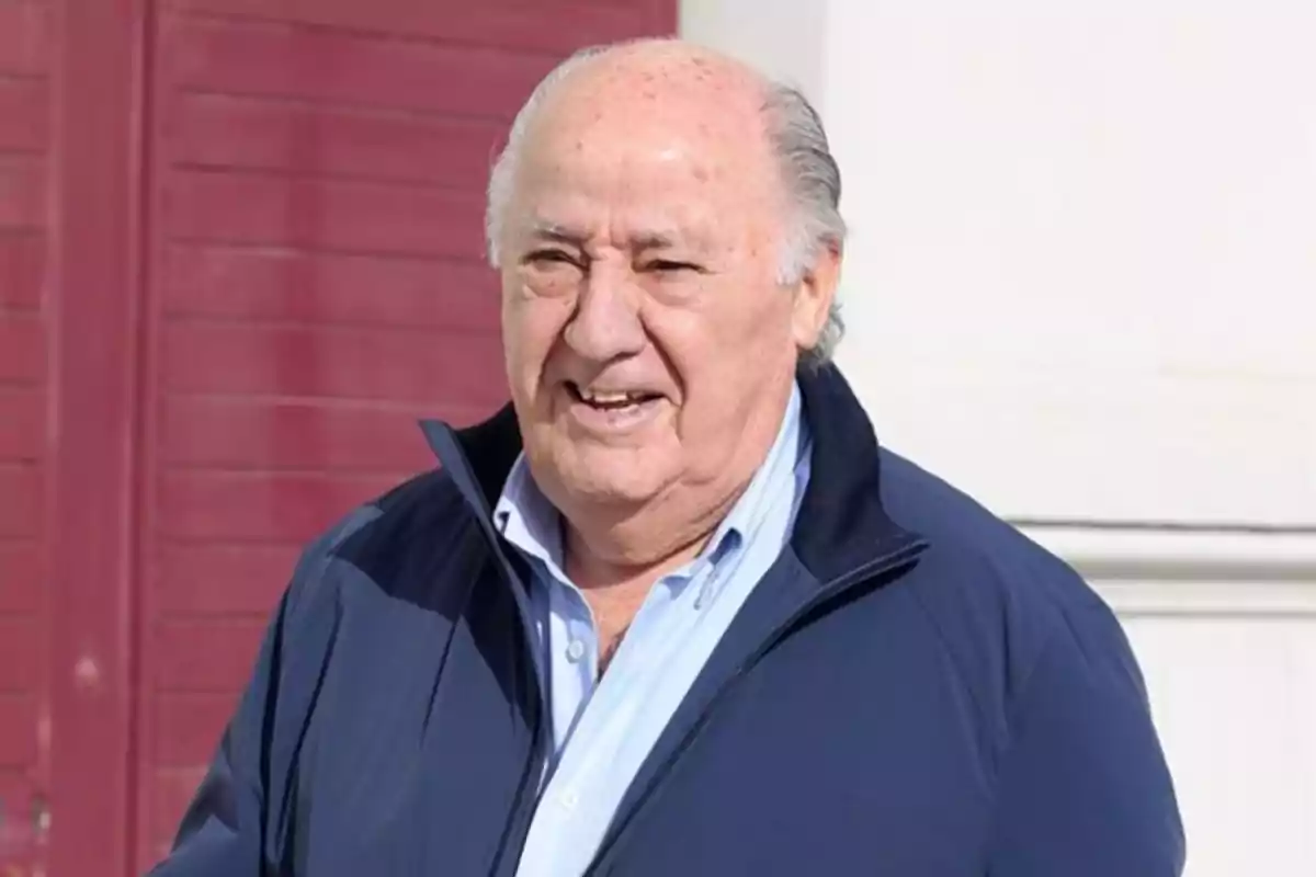 Amancio Ortega con chaqueta azul y camisa clara sonríe mientras está de pie frente a una pared de color rojo oscuro. Amancio Ortega con chaqueta azul y camisa clara sonríe mientras está de pie frente a una pared de color rojo oscuro.
