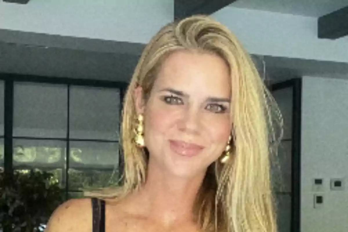 Una persona con cabello rubio y aretes dorados, sonriendo en un ambiente interior con ventanas grandes al fondo. Una persona con cabello rubio y aretes dorados, sonriendo en un ambiente interior con ventanas grandes al fondo.