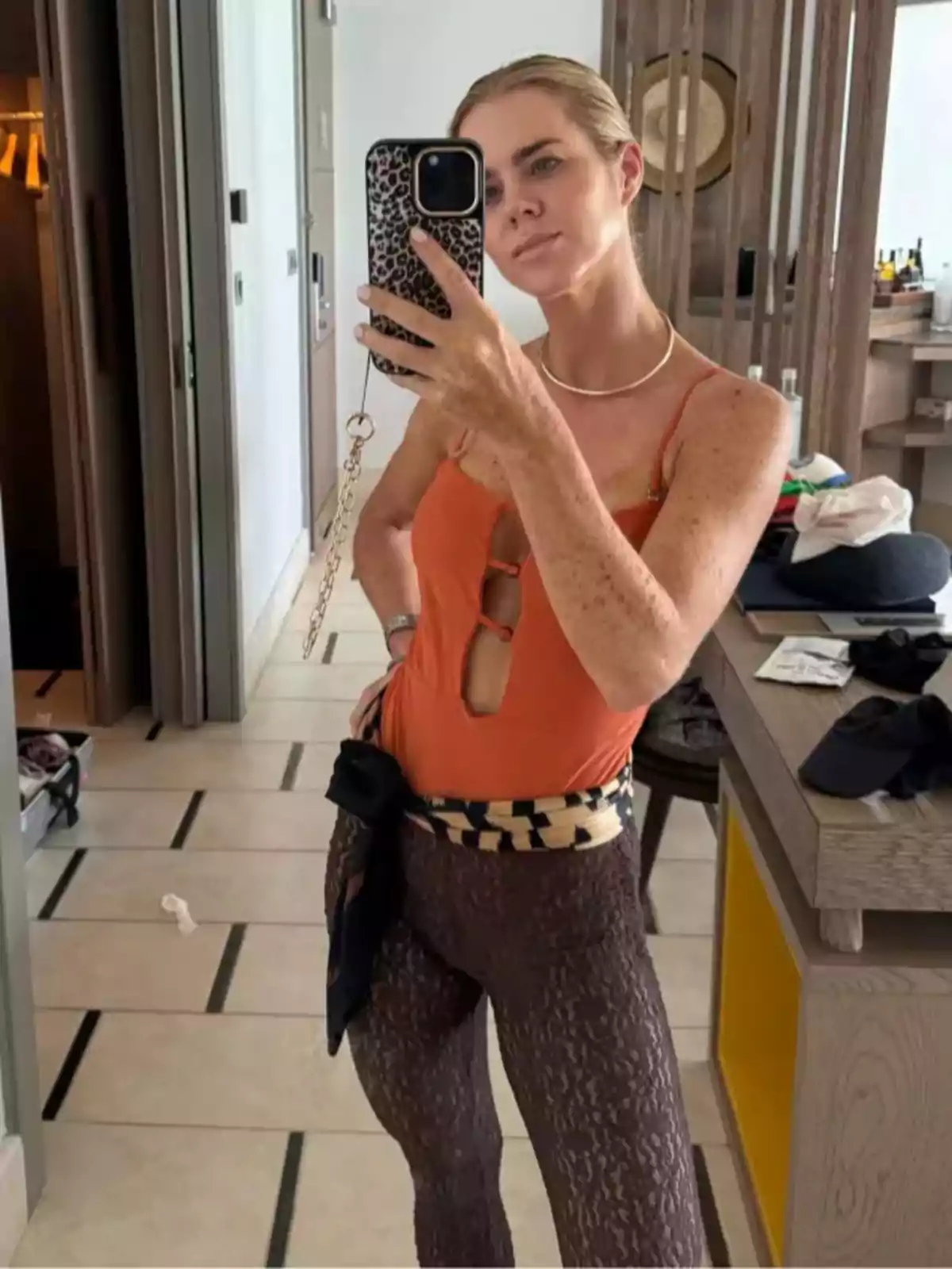 Persona tomándose una selfie frente a un espejo, vistiendo un traje de baño naranja y pantalones de encaje marrón, con un teléfono de carcasa de leopardo. Persona tomándose una selfie frente a un espejo, vistiendo un traje de baño naranja y pantalones de encaje marrón, con un teléfono de carcasa de leopardo.