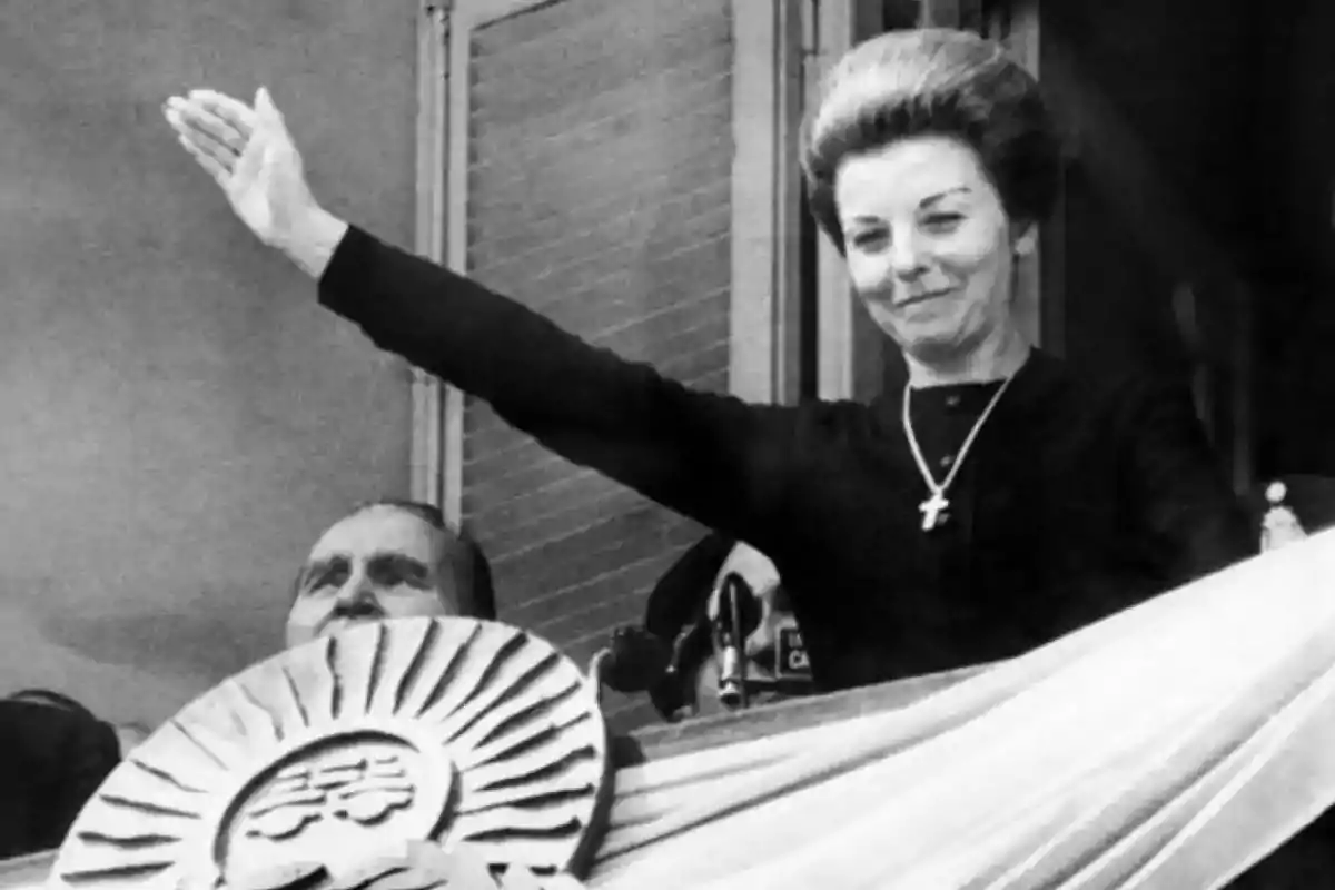 Una mujer con un vestido oscuro y un collar con una cruz está saludando desde un balcón, mientras un hombre está parcialmente visible detrás de un adorno decorativo.