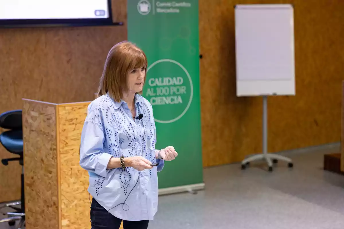 Una mujer dando una presentación en un entorno profesional con un cartel verde que dice "Calidad al 100 por Ciencia" y un rotafolio en el fondo.