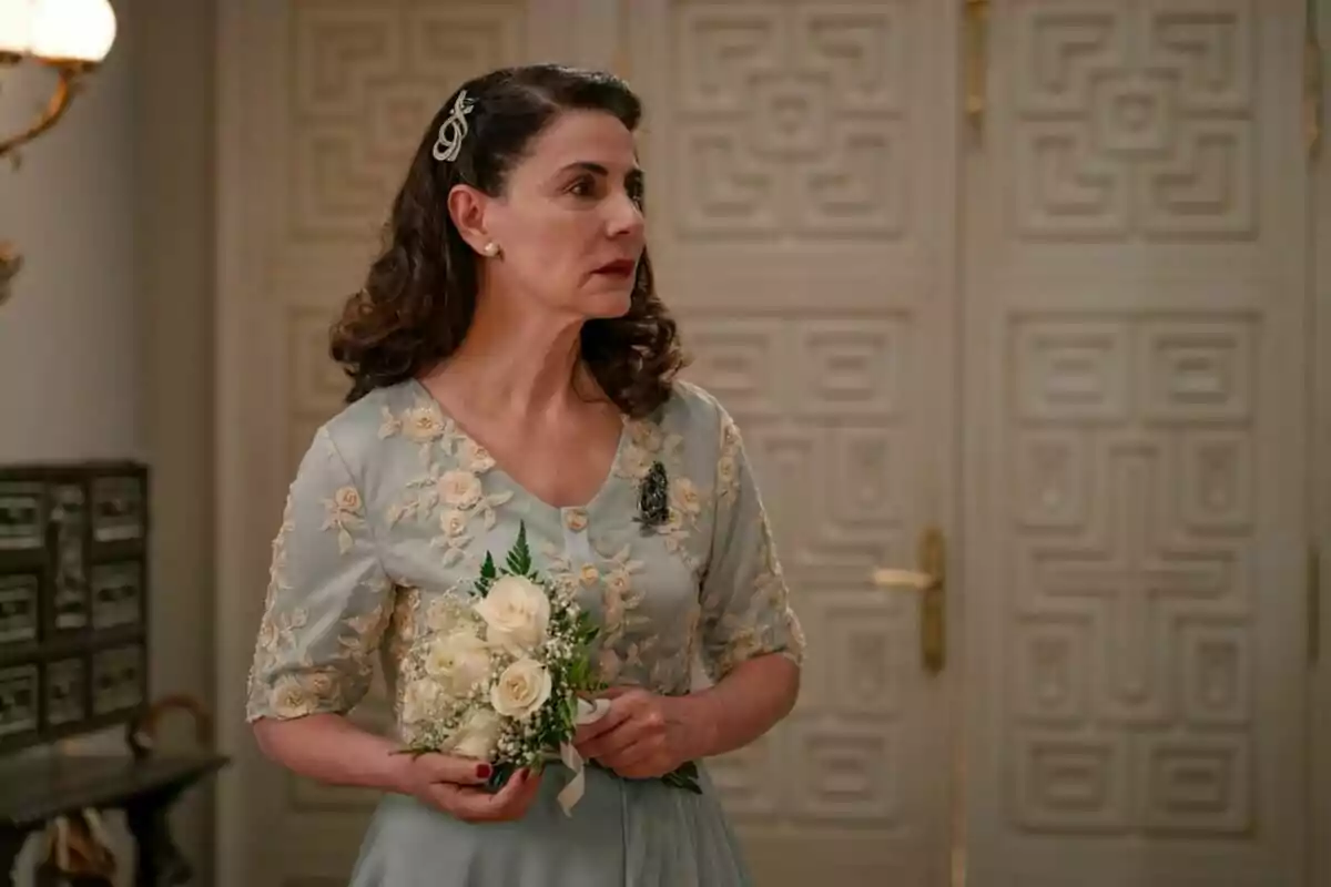 La actriz Ana Fernández en una escena de 'Sueños de Libertad' con vestido azul claro bordado sosteniendo un pequeño ramo de flores blancas en un interior elegante. La actriz Ana Fernández en una escena de 'Sueños de Libertad' con vestido azul claro bordado sosteniendo un pequeño ramo de flores blancas en un interior elegante.