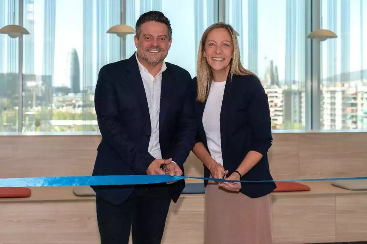 Dos personas sonrientes cortando una cinta azul en una ceremonia de inauguración con un fondo de ventanas grandes y una vista urbana.