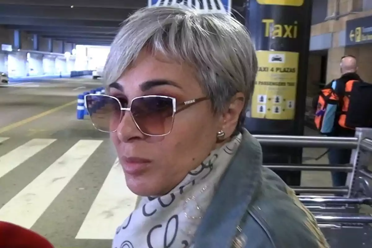 Ana María Aldón con gafas de sol y cabello corto canoso está en un área de taxis. Ana María Aldón con gafas de sol y cabello corto canoso está en un área de taxis.