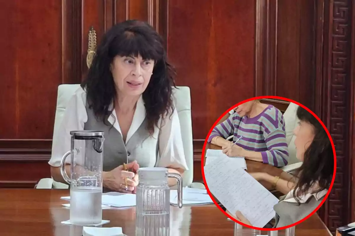 Una mujer con cabello oscuro y vestimenta formal está sentada en una mesa de madera con una jarra de agua y papeles, mientras en un recuadro se observa a otra persona escribiendo en un cuaderno.