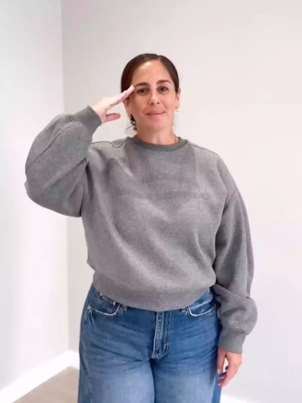 Una persona con suéter gris y jeans azules hace un saludo militar mientras sonríe. Una persona con suéter gris y jeans azules hace un saludo militar mientras sonríe.
