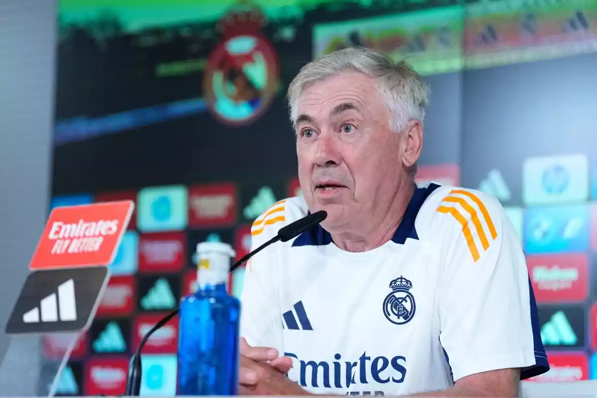 Un hombre de cabello canoso y camiseta blanca del Real Madrid está hablando en una conferencia de prensa, con un micrófono frente a él y un fondo de logotipos de patrocinadores.