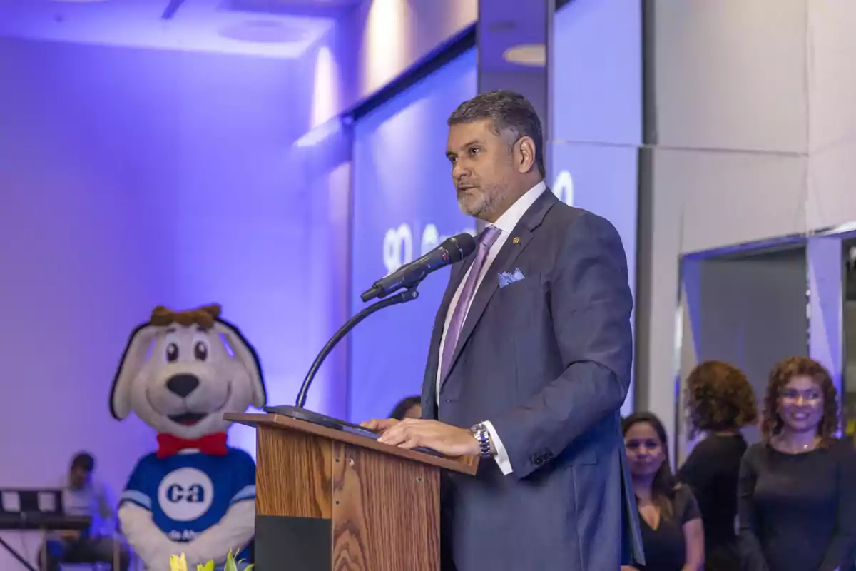 Hombre de traje hablando en un podio durante un evento formal con una mascota de perro gigante al fondo y varias personas observando