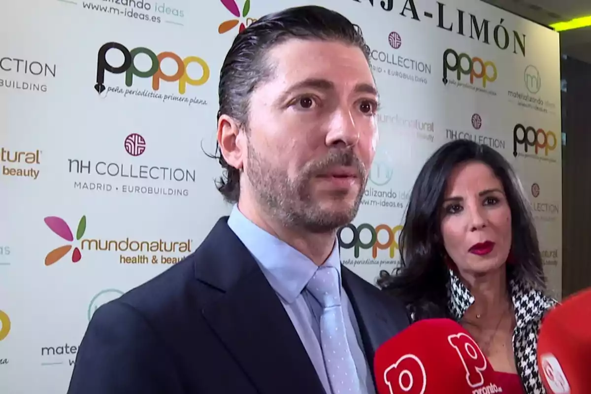 Ángel Cristo con traje y corbata es entrevistado por la prensa mientras Ana Herminia con labios rojos lo acompaña, ambos están frente a un fondo con logotipos de diferentes marcas y medios. Ángel Cristo con traje y corbata es entrevistado por la prensa mientras Ana Herminia con labios rojos lo acompaña, ambos están frente a un fondo con logotipos de diferentes marcas y medios.