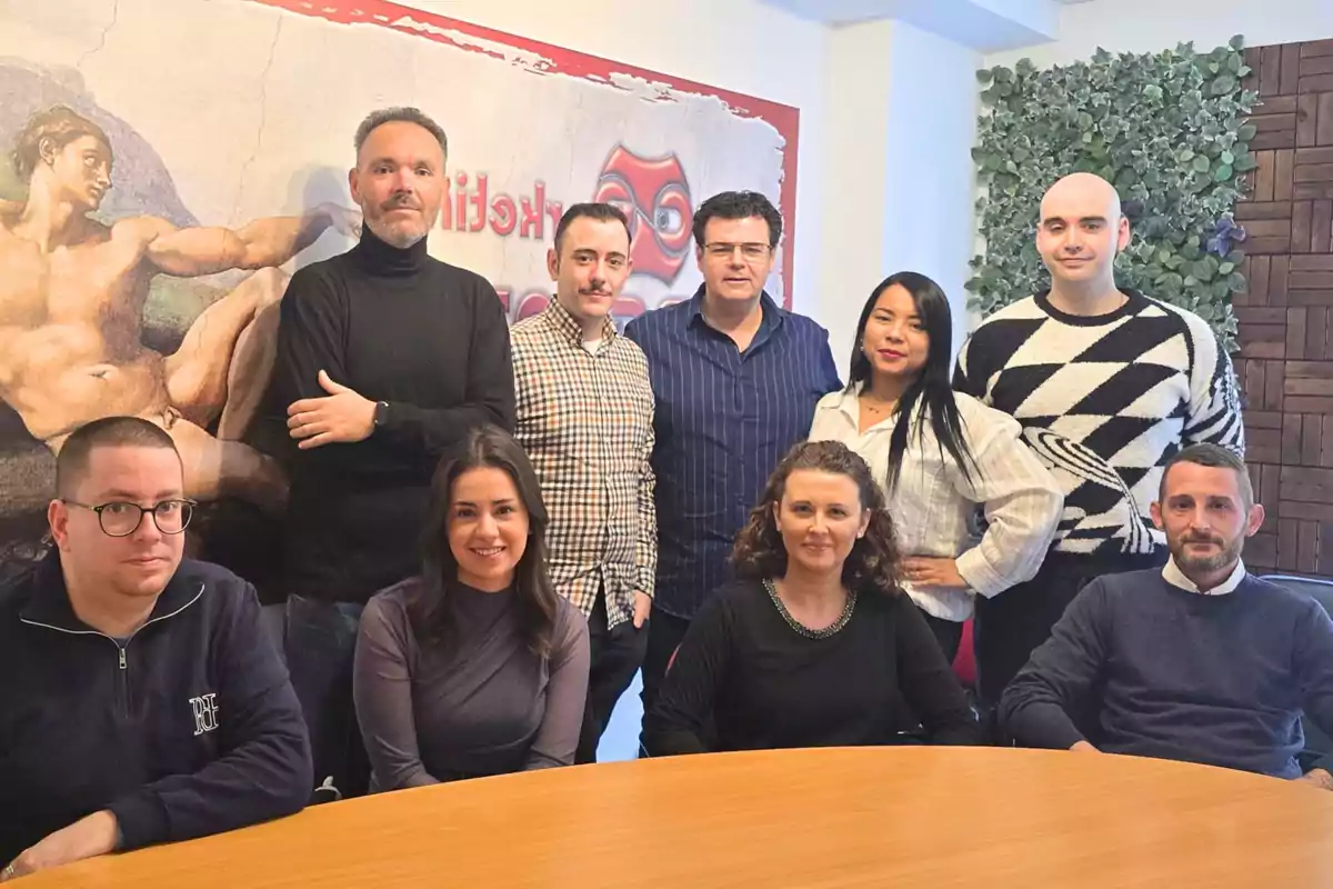 Un grupo de personas posando juntas en una sala de reuniones con una mesa de madera en primer plano y un mural artístico en la pared de fondo.
