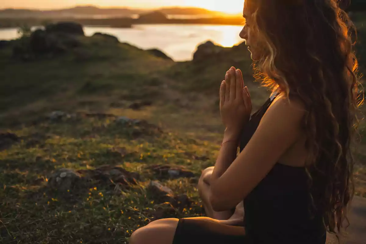 Mujer meditando al aire libre al atardecer con las manos juntas y el sol brillando en el fondo.