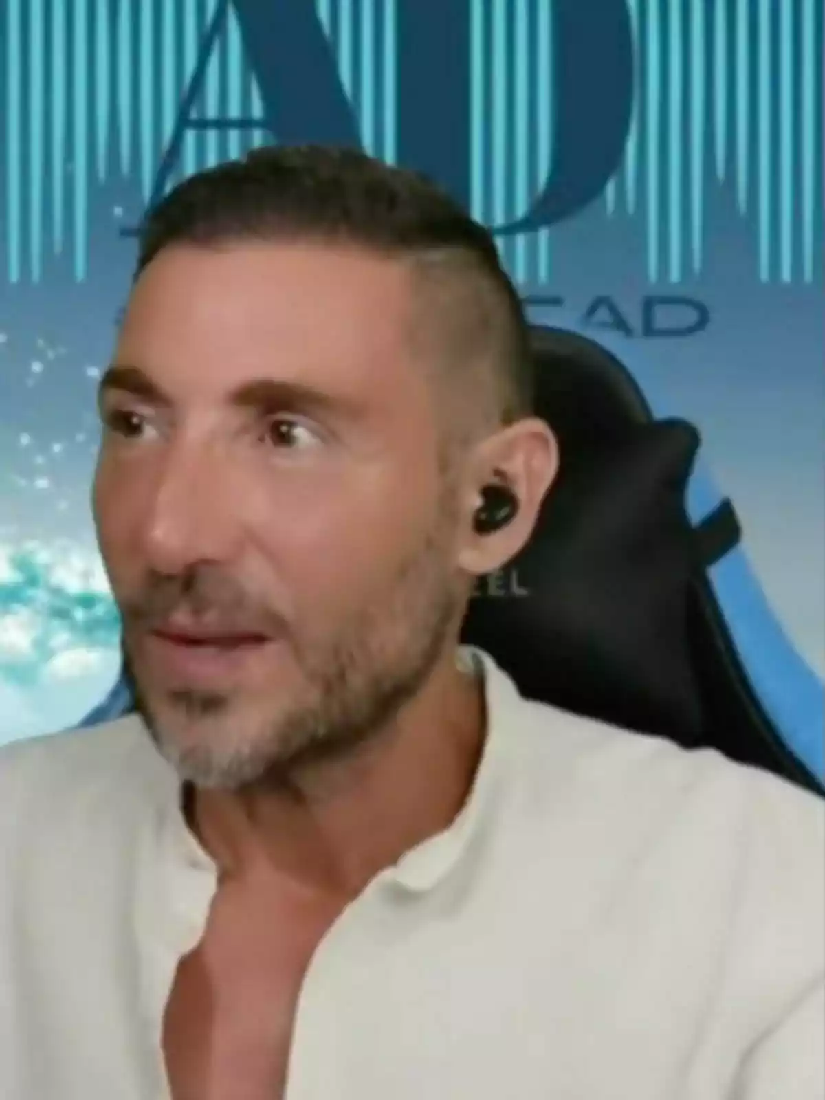 Antonio David Flores con auriculares negros y camisa blanca, sentado frente a un fondo azul con letras. Antonio David Flores con auriculares negros y camisa blanca, sentado frente a un fondo azul con letras.