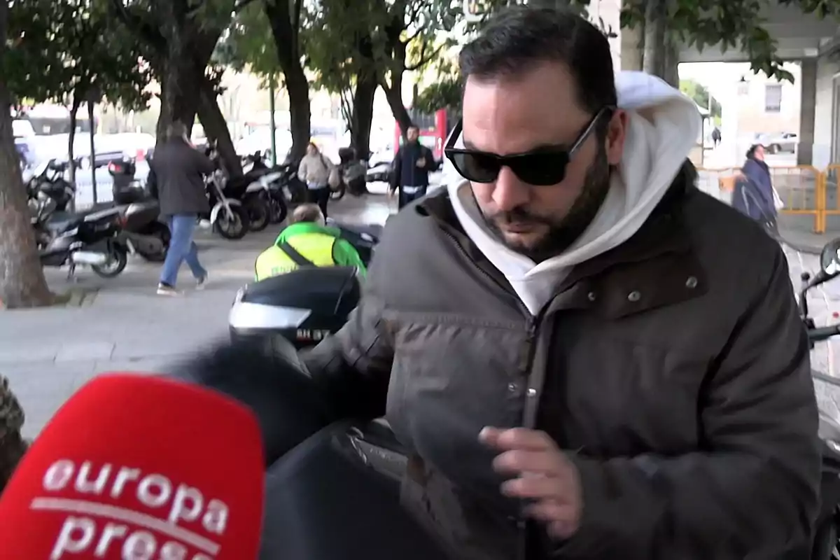 Antonio Tejado wearing sunglasses and a brown jacket walks down the street while a Europa Press microphone appears in the foreground.