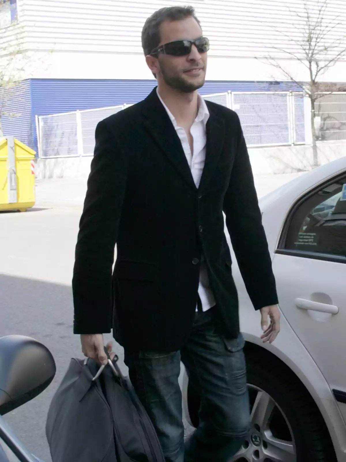 Antonio Tejado con gafas de sol, chaqueta negra y camisa blanca lleva una bolsa mientras camina junto a un coche en un entorno urbano. Antonio Tejado con gafas de sol, chaqueta negra y camisa blanca lleva una bolsa mientras camina junto a un coche en un entorno urbano.