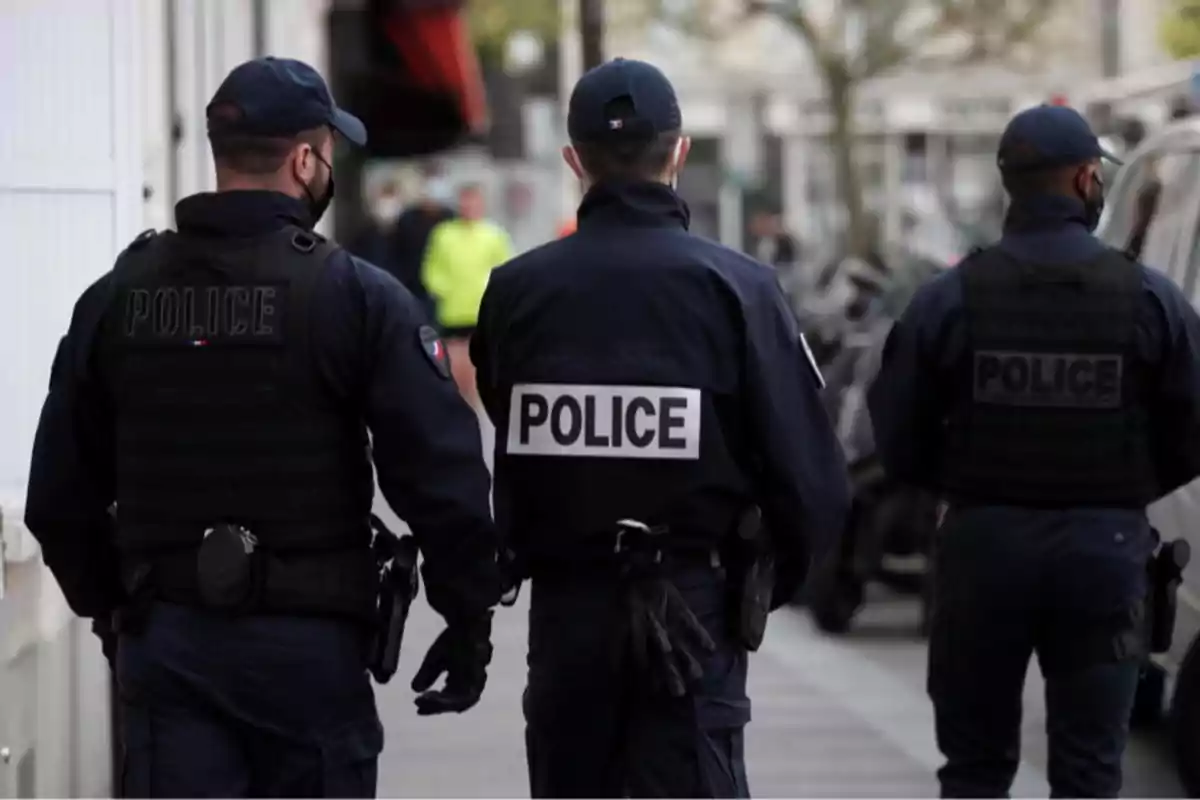 Tres policías de espaldas caminan por una acera vestidos con uniforme azul oscuro y chalecos antibalas Tres policías de espaldas caminan por una acera vestidos con uniforme azul oscuro y chalecos antibalas